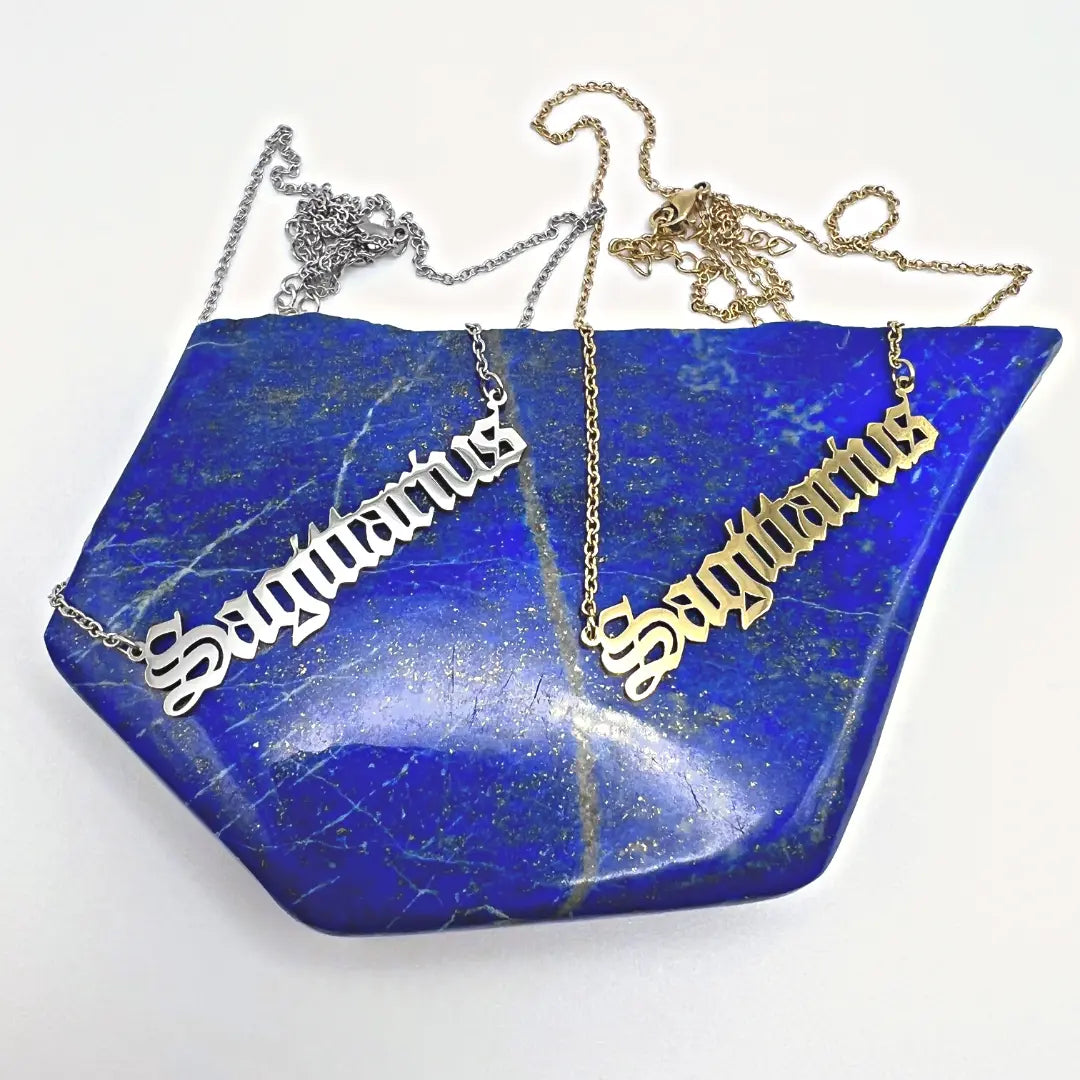 Ascention Beauty's Zodiac Necklaces in gold and silver are delicately placed on a textured blue stone background, featuring the Sagittarius sign and embodying a celestial identity.