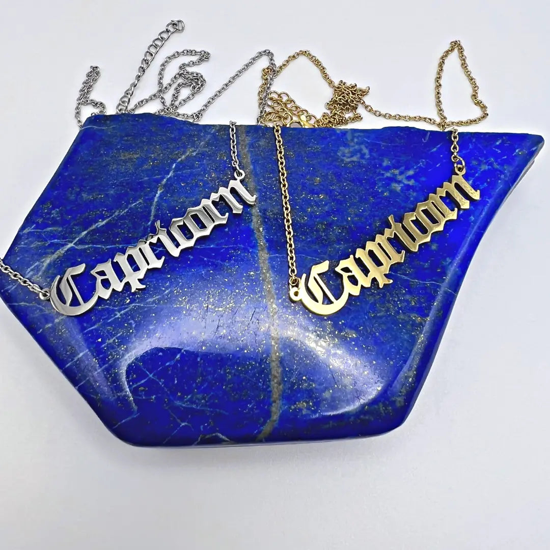 Two Zodiac Necklaces elegantly display the word "Capricorn" in cursive on both gold and silver chains. These exquisite pieces, from Ascention Beauty's collection of Wellness Clean Cruelty-Free Vegan Perfume, rest against a blue, gemstone-like surface adorned with gold flecks, beautifully capturing a celestial identity.
