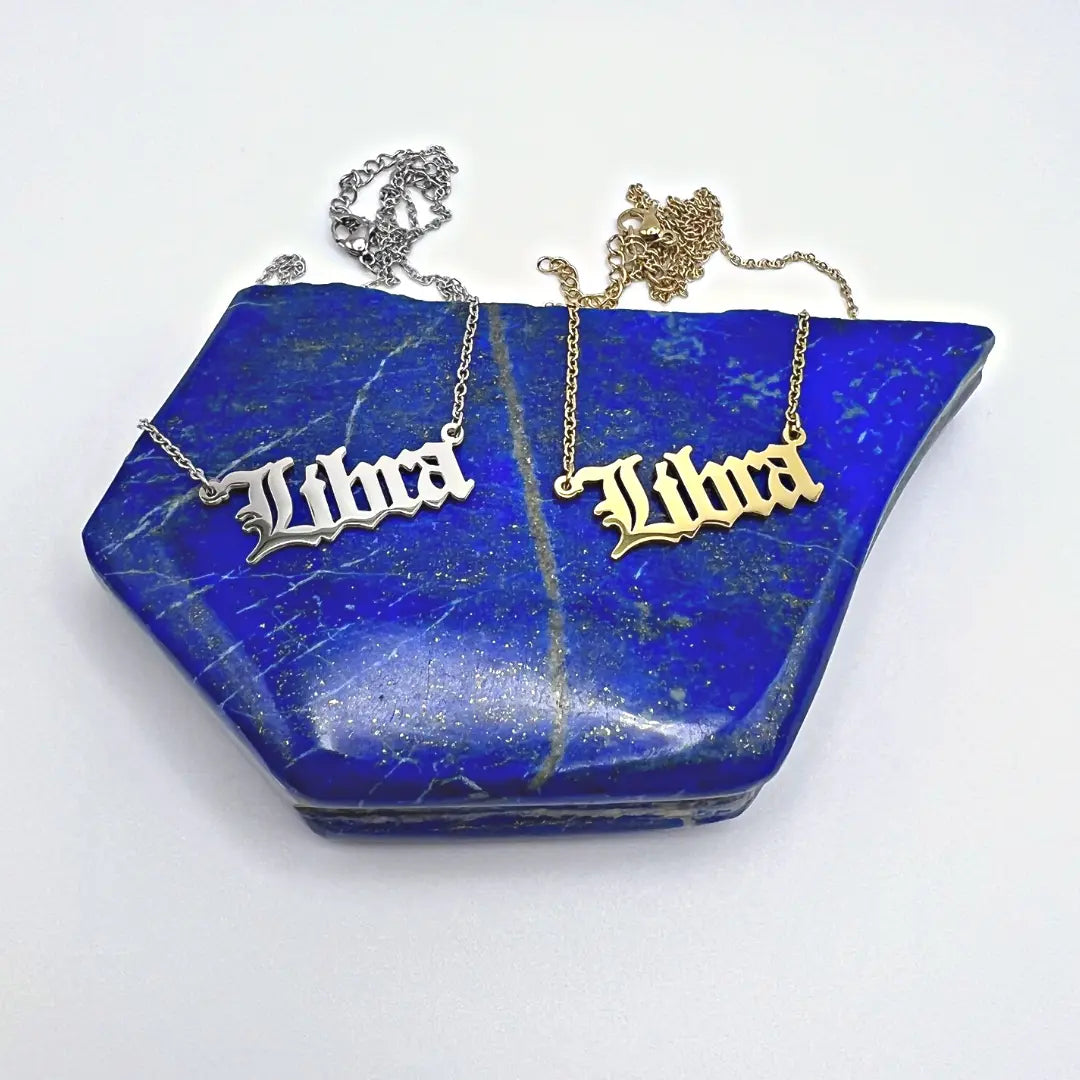Displayed on a textured blue stone with golden flecks, the Ascention Beauty Zodiac Necklaces feature silver and gold Libra nameplates. These necklaces capture a celestial identity and cosmic essence, embodying elegance and astrological charm against a plain white background.