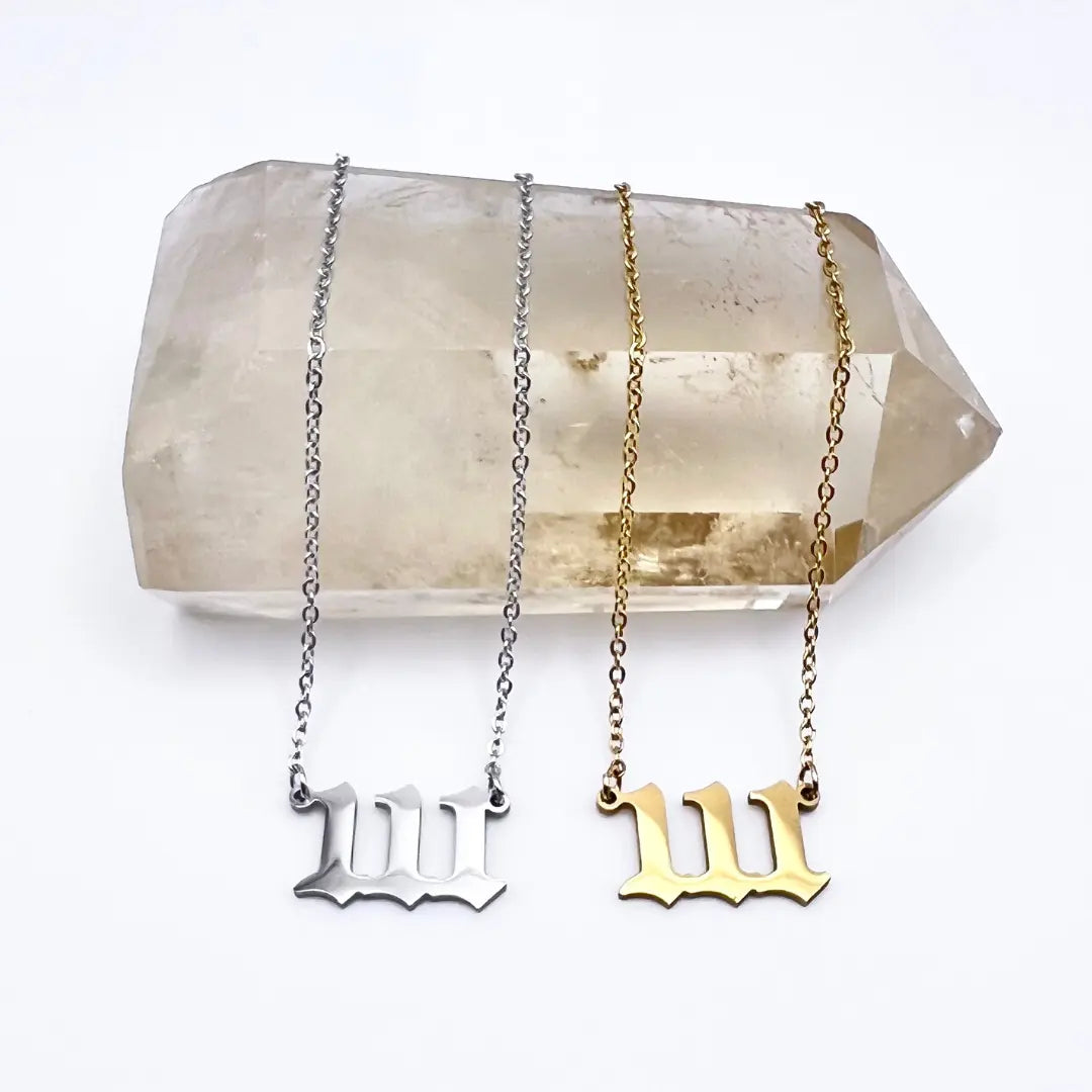 Two Angel Number Necklaces by Ascention Beauty - Wellness Clean Cruelty-Free Vegan Perfume, featuring chain links and M-shaped pendants in silver and gold, are elegantly displayed on a clear quartz crystal known for amplifying spiritual guidance. The display rests against a plain white backdrop.