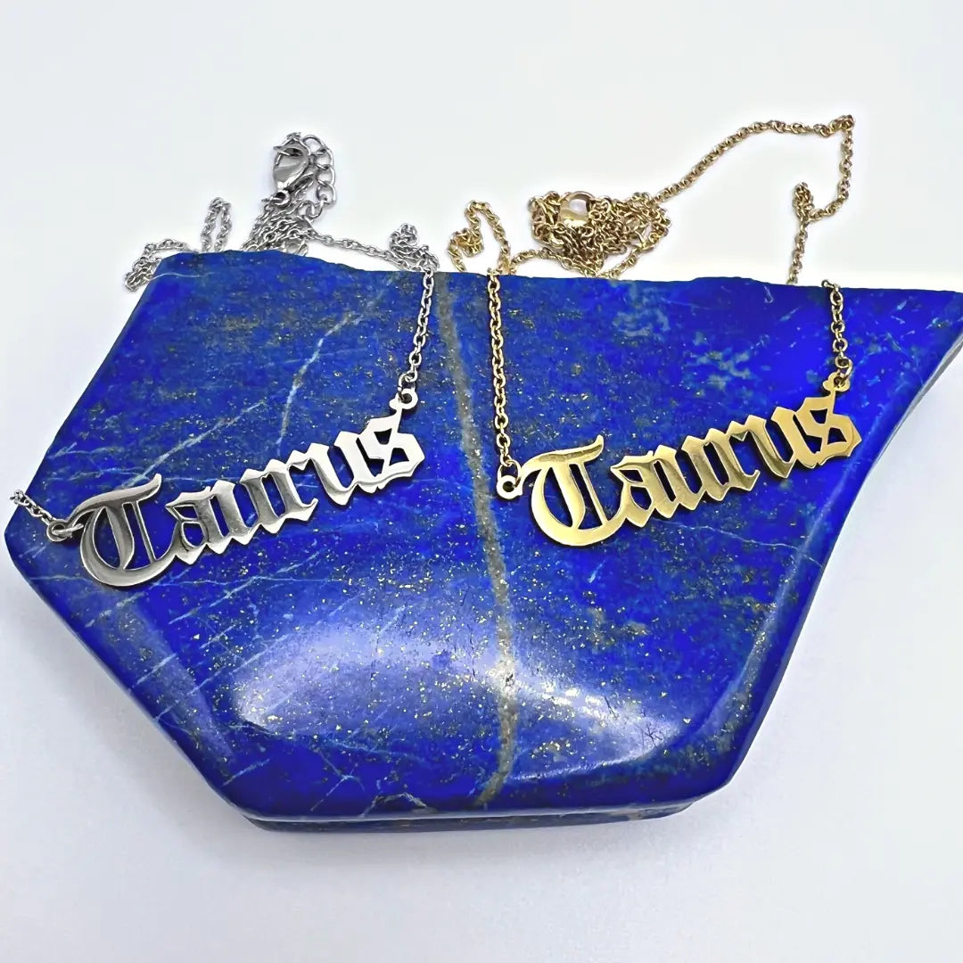Two Zodiac Necklaces from Ascention Beauty, featuring Taurus pendants that exude a cosmic charm in silver and gold, are gracefully displayed on a polished blue stone slab accented with golden specks.