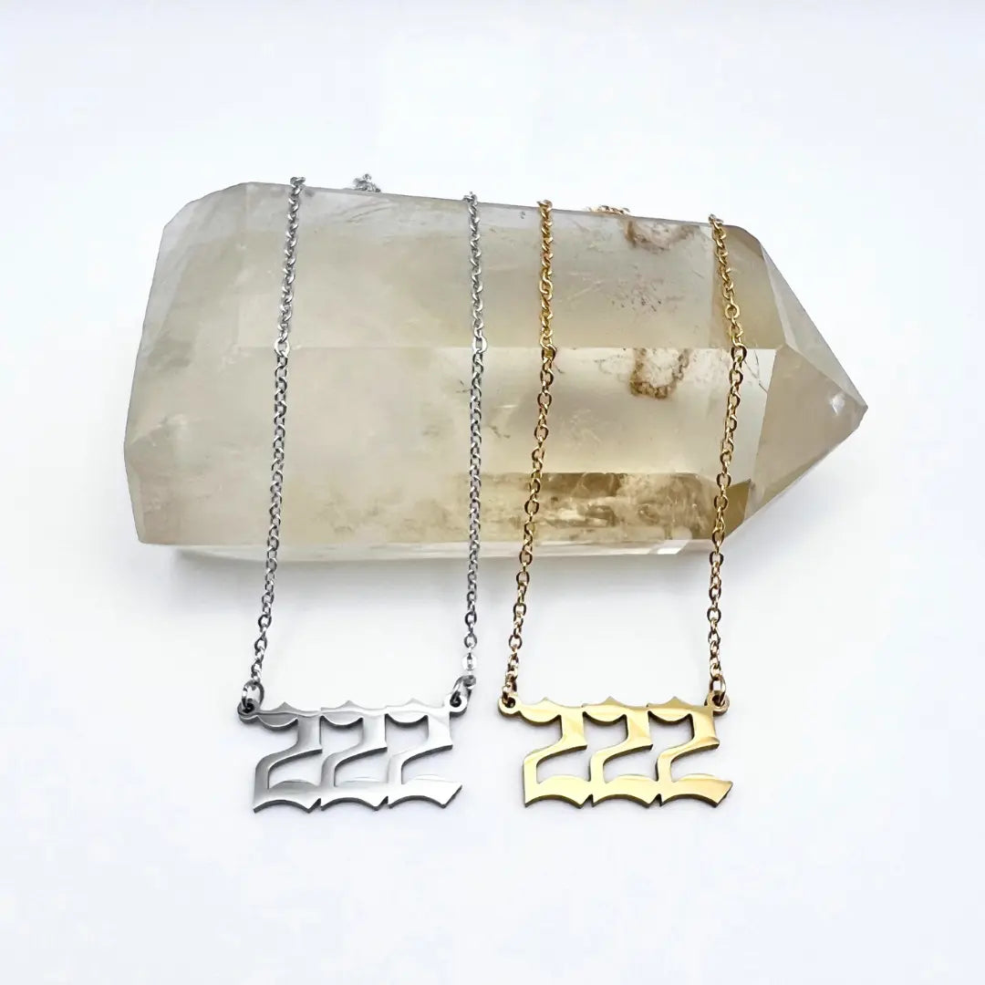 Two Angel Number Necklaces by Ascention Beauty are elegantly displayed on a large clear quartz crystal against a white background. Each necklace features a chain pendant shaped as 222, available in shimmering metallic shades of silver and gold. These pieces are perfect for those seeking spiritual guidance and manifesting desires, bringing together beauty and intention from this wellness-focused, clean, cruelty-free brand.