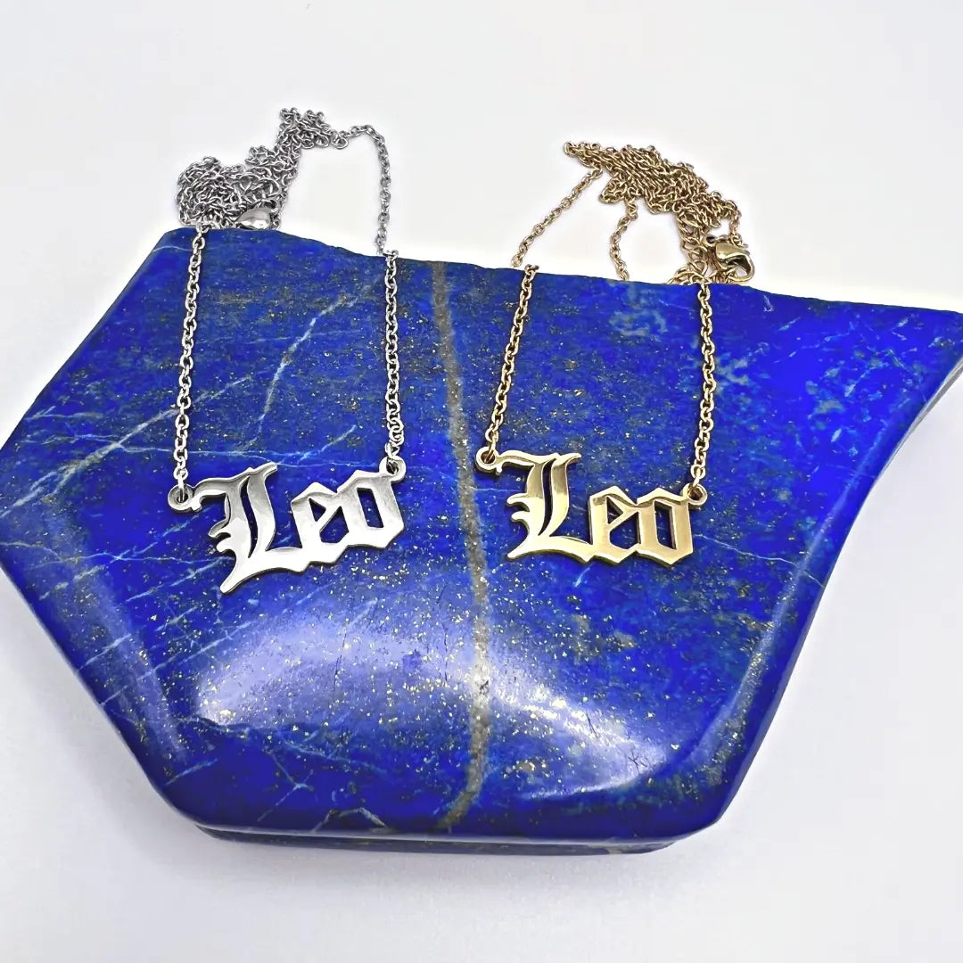 Two Zodiac Necklaces from Ascention Beauty - Wellness Clean Cruelty-Free Vegan Perfume, featuring Leo pendants in silver and gold, capture a celestial identity as they rest on a polished blue stone surface. The plain white background enhances their cosmic essence.