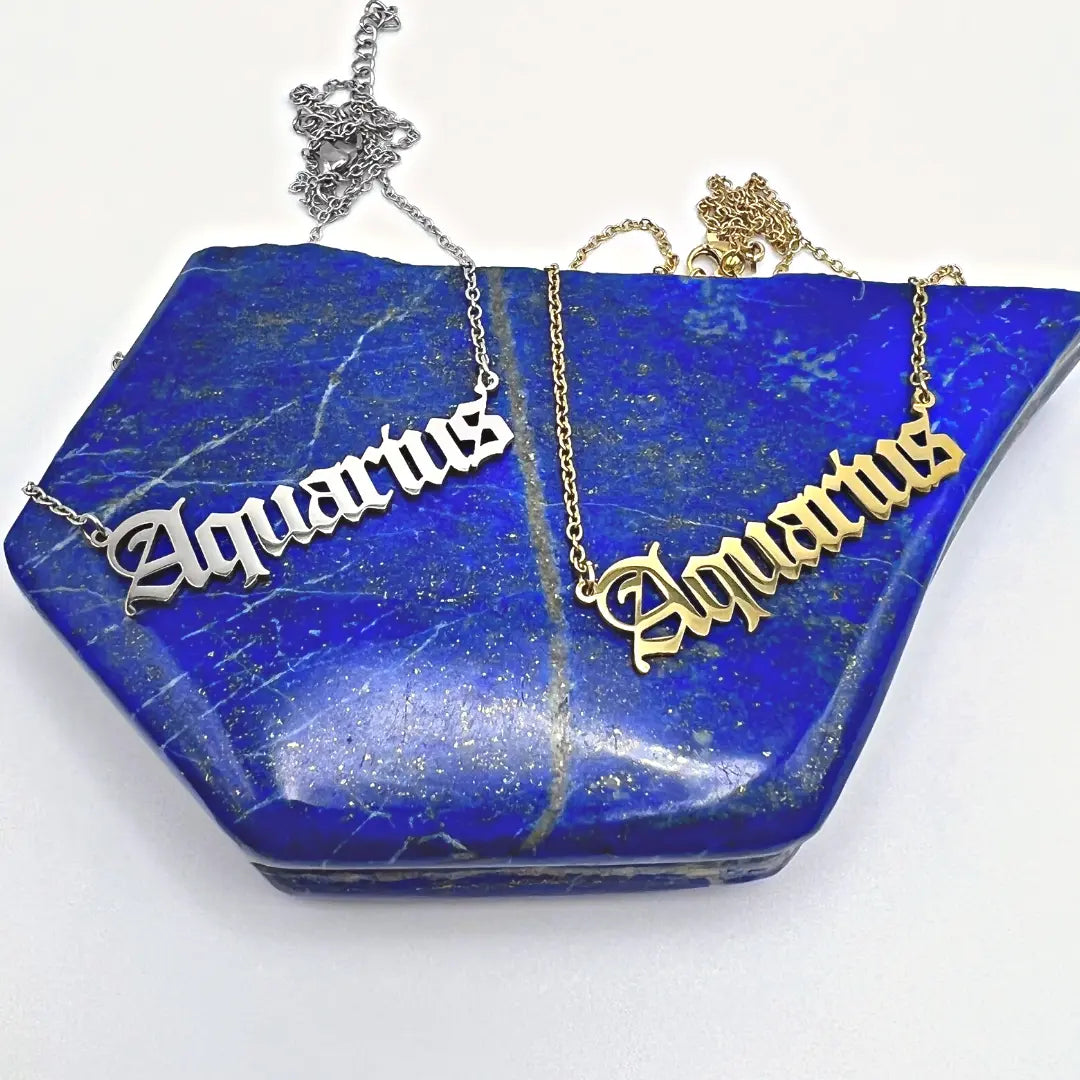 Two exquisite Zodiac Necklaces from Ascention Beauty are elegantly displayed on a lapis lazuli stone, showcasing the celestial identity of their wearer with the word Aquarius. One necklace is crafted in silver and the other in gold, both featuring a stylish script font to embody your cosmic essence.