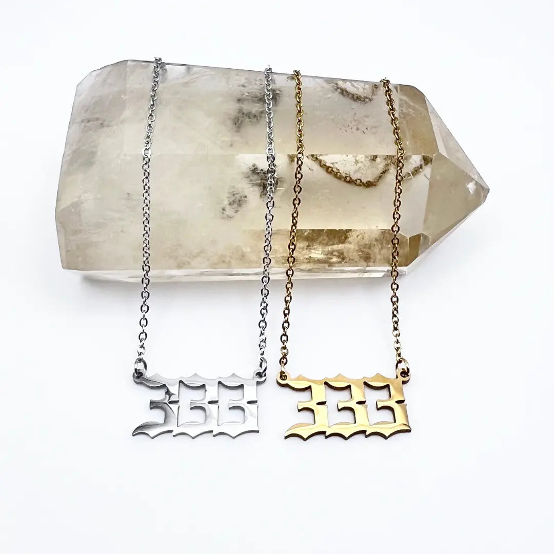 Two *Angel Number Necklaces* from *Ascention Beauty - Wellness Clean Cruelty-Free Vegan Perfume* are displayed on a large clear crystal with natural inclusions, representing angel messages. One necklace features a silver pendant, and the other a gold one, both designed to capture whispers of divine guidance. A white background enhances their minimalist presentation.