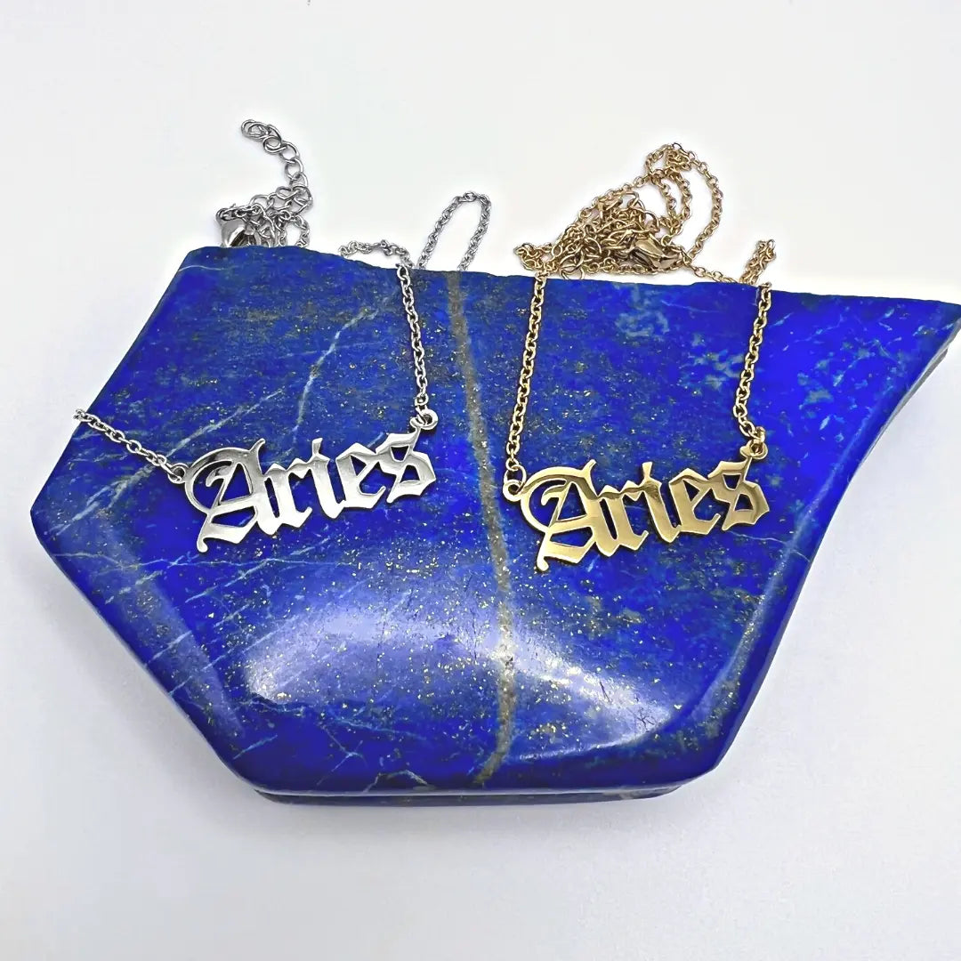 Displayed on a blue, angular stone surface, two Zodiac Necklaces with Aries pendants in silver and gold from Ascention Beauty - Wellness Clean Cruelty-Free Vegan Perfume beautifully capture a celestial identity. The chain of each necklace is artfully laid out above their matching pendants.