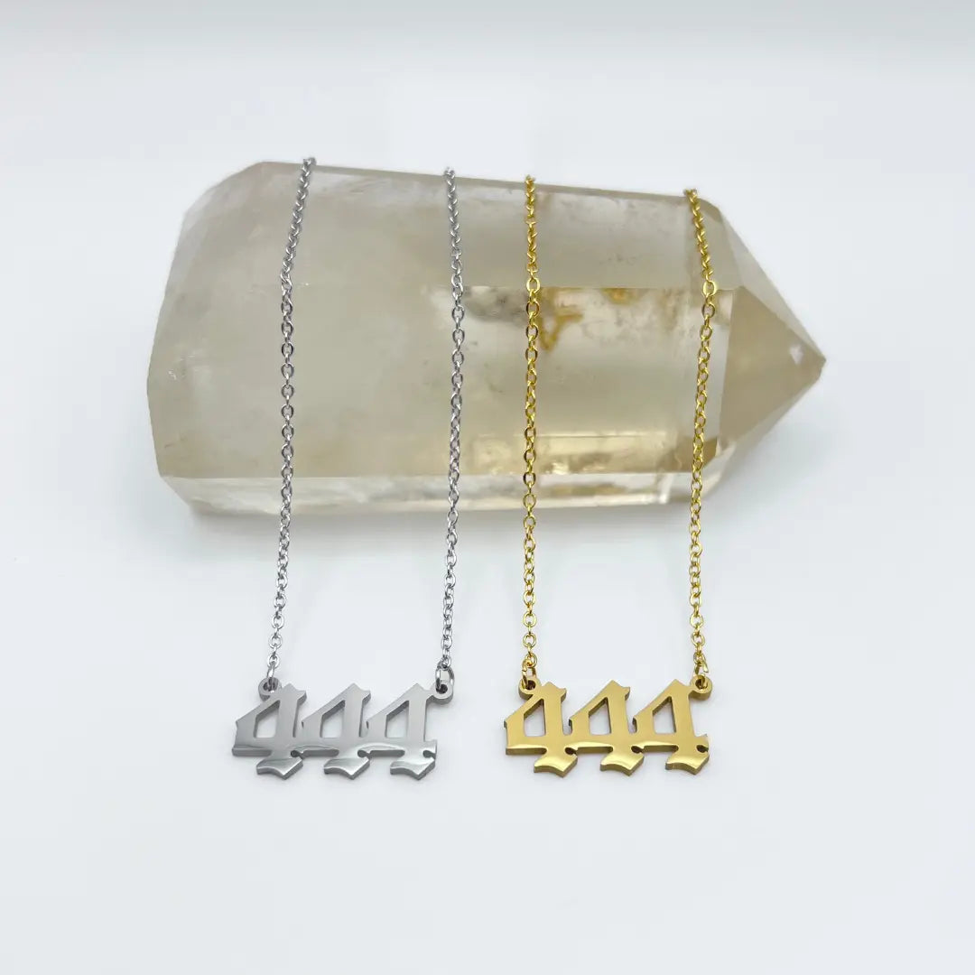 Two Angel Number Necklaces from Ascention Beauty are elegantly displayed on a clear quartz crystal. Each necklace features a pendant with the number 444, representing angelic messages. One is crafted in silver, and the other in gold, both exquisitely showcased against a simple white background to invite spiritual guides to manifest desires.