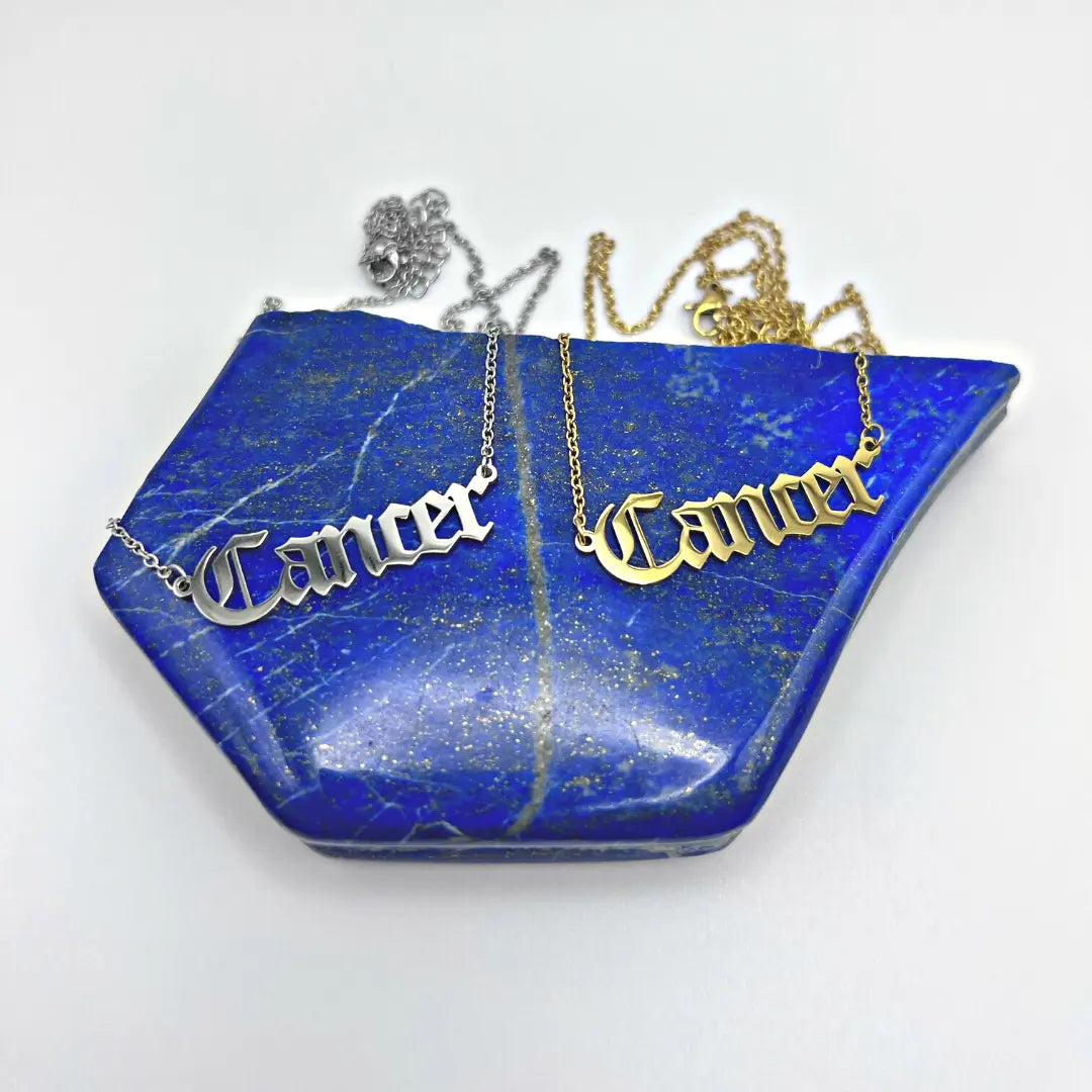 A pair of Zodiac Necklaces by Ascention Beauty—featuring the word "Cancer" in Gothic font—gleam in silver and gold atop a blue stone sprinkled with gold specks, capturing a celestial essence.