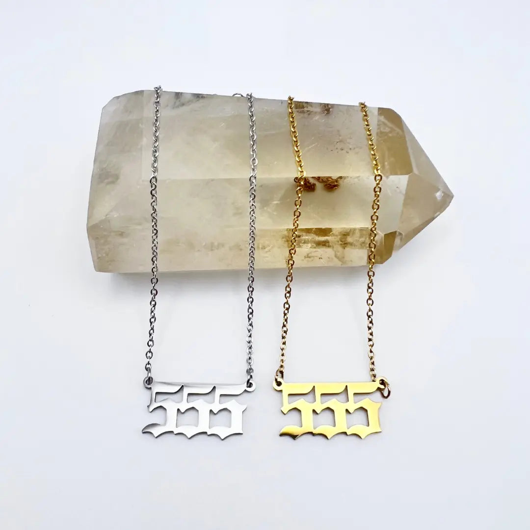 Two Angel Number Necklaces from Ascention Beauty are displayed on a large, clear quartz crystal, symbolizing divine guidance. One necklace features a silver design, while the other is crafted in gold. They rest against a plain white surface, which enhances their spiritual significance.