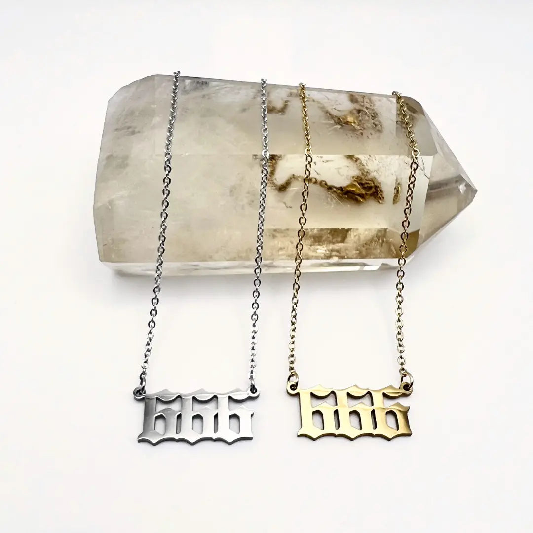 The Angel Number Necklaces from Ascention Beauty elegantly drape over a large, clear quartz crystal, embodying divine guidance. Each necklace—one in silver and the other in gold—features intricate designs with 666 pendants. Against the white background, the distinct contrast between the metals and the crystal creates a mystical aura that enhances this spiritual piece.
