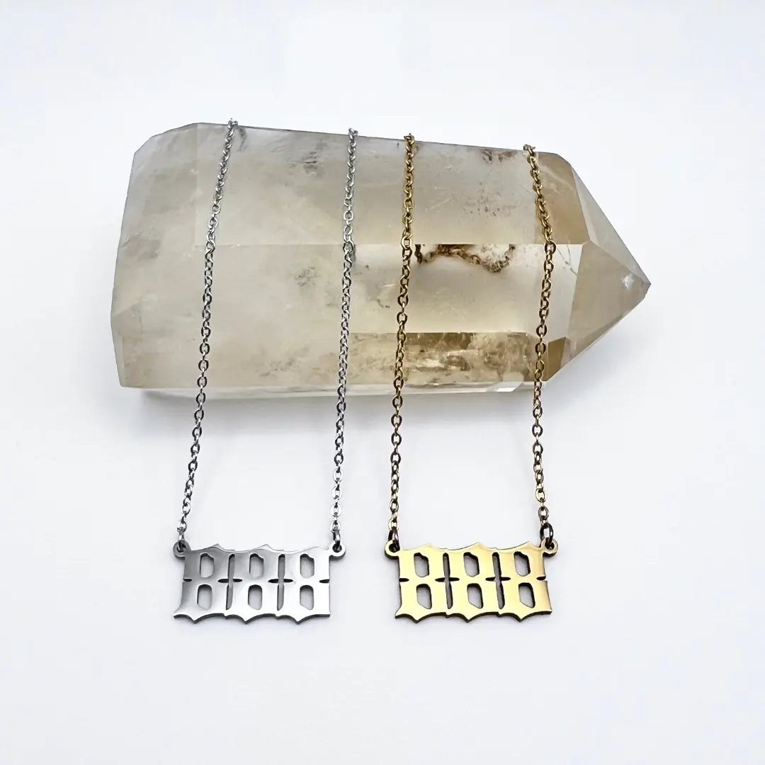 Displayed on a large, translucent crystal are two Angel Number Necklaces from Ascention Beauty - Wellness Clean Cruelty-Free Vegan Perfume. A silver and a gold necklace, both featuring stylized number pendants that resonate with angel messages, gracefully dangle on delicate chains, offering whispers of divine guidance.