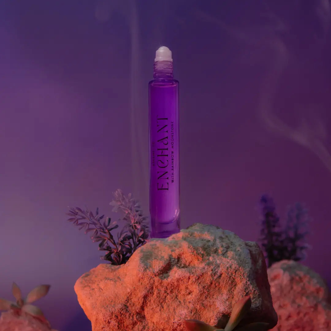 A purple perfume bottle labeled as "*NEW* Ascent to Enchant With Rainbow Moonstone Perfume" by ASCENTION BEAUTY CO stands upright on a rock, surrounded by small plants. Set against a dreamy, smoky purple and pink background, it encapsulates the essence of a mystical fragrance inspired by the dark feminine goddess.